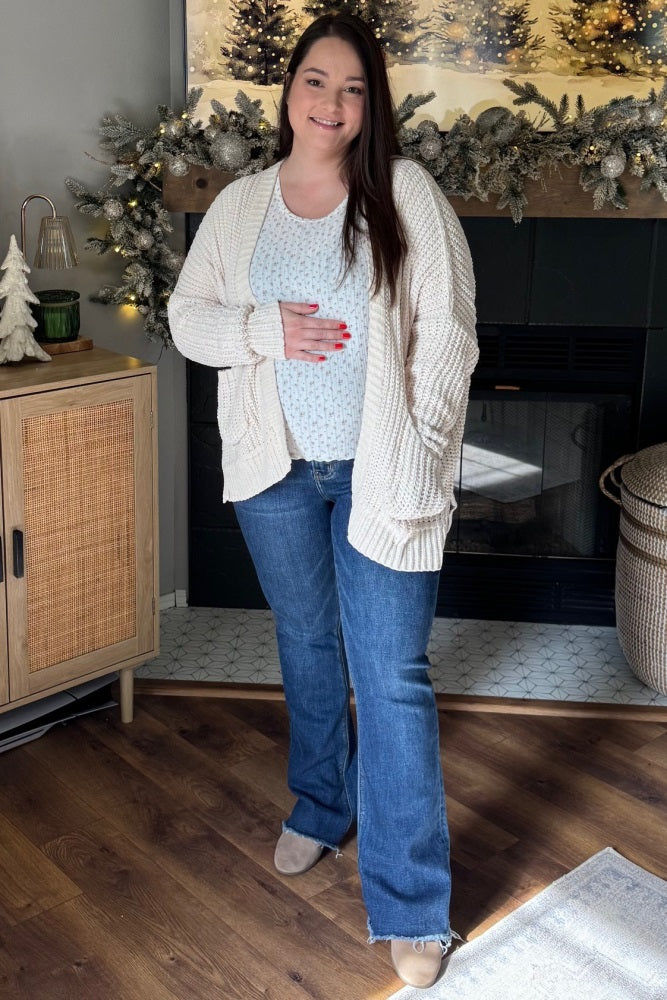 woman wearing a cream chunky knit cardigan and blue maternity top, paired with flared jeans, holding her baby bump