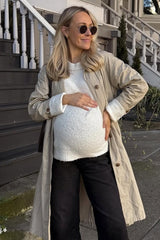 woman wearing a beige trench coat, white knit maternity top, and black pants, showing her baby bump