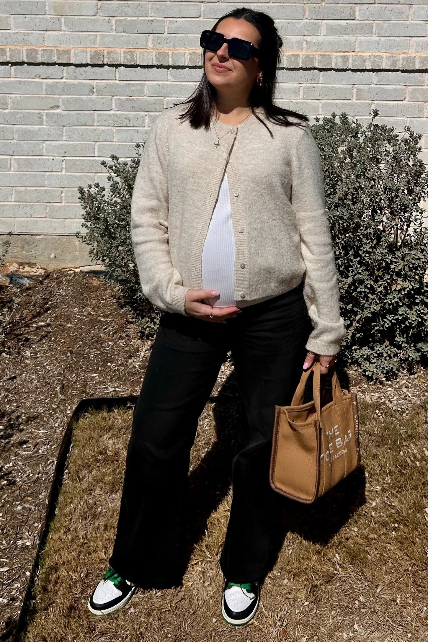 woman wearing a cream cardigan over a white maternity top, holding a brown tote bag and showing her baby bump