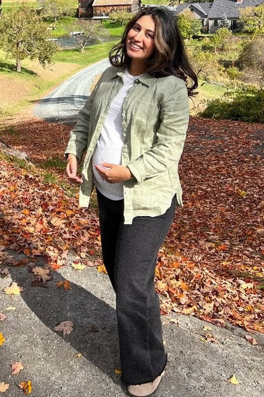 woman wearing a light green maternity shirt jacket and white top, holding her baby bump, standing on a path with autumn leaves