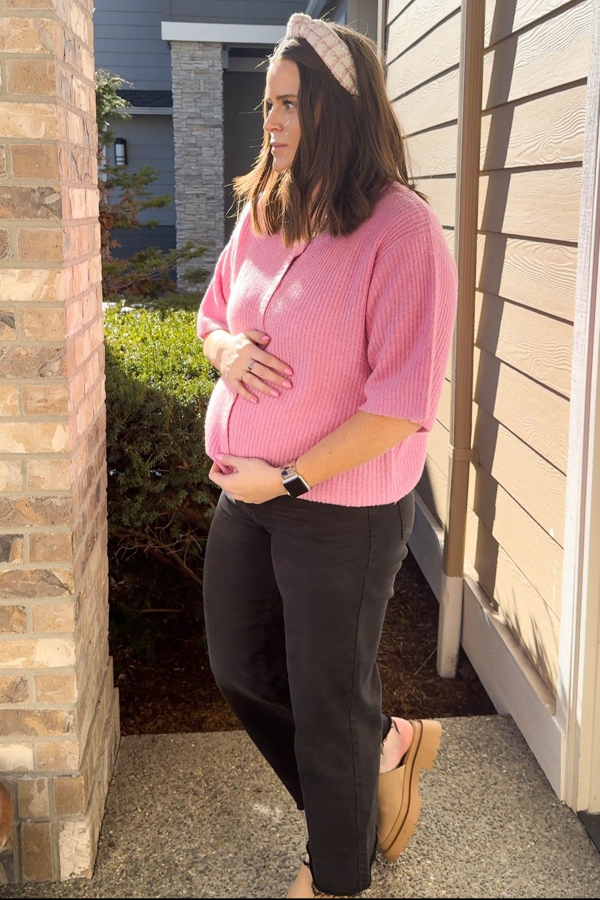 woman wearing a pink short sleeve maternity sweater and black pants, holding her baby bump outside