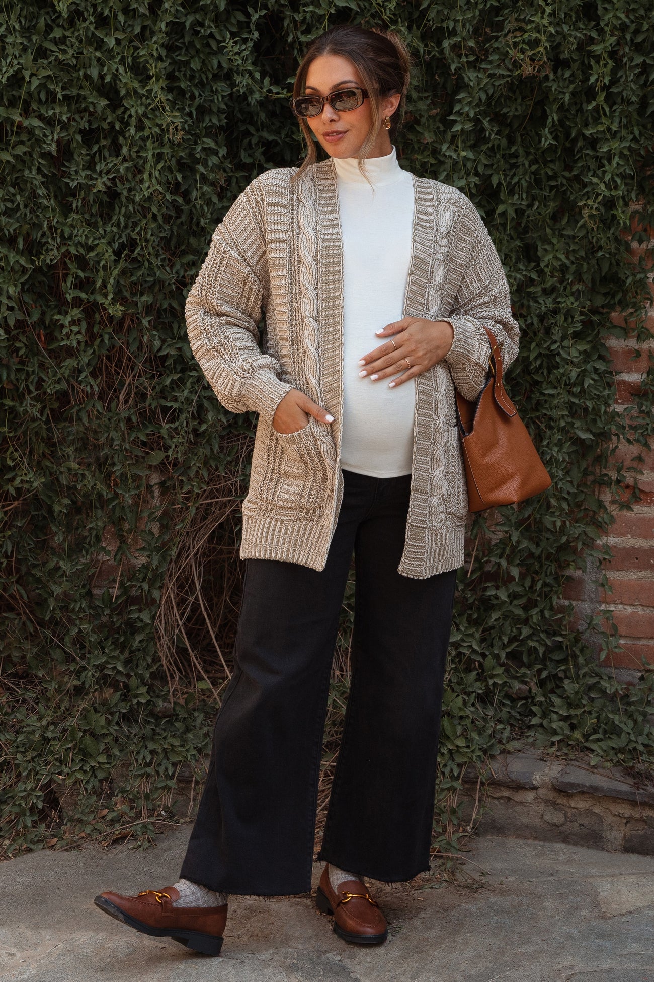 woman wearing a beige knitted maternity cardigan over a white top and black wide-leg pants, holding a brown bag and showing her baby bump