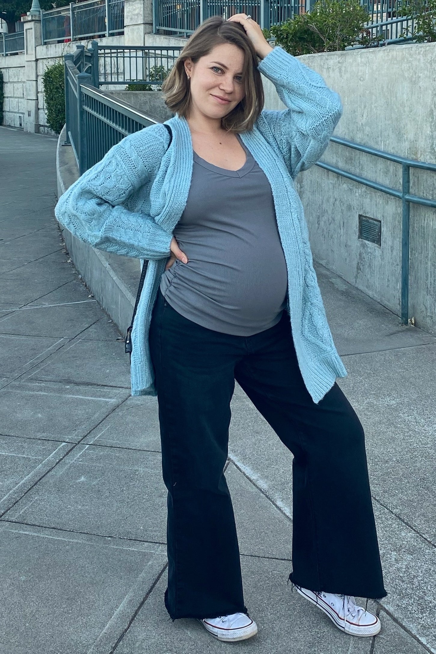 woman wearing a light blue cable-knit cardigan over a gray maternity top, posing with a visible baby bump and black flared pants