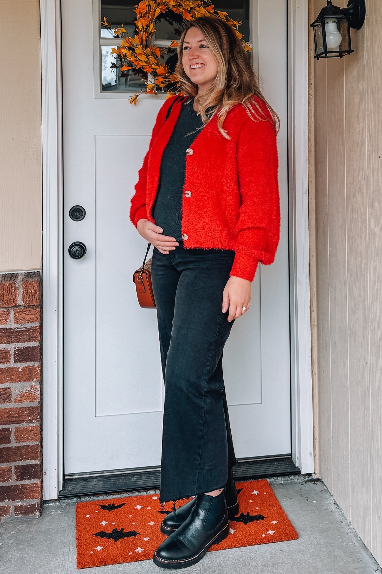 woman wearing a bright red fluffy maternity cardigan and black flared jeans, holding her baby bump by the front door