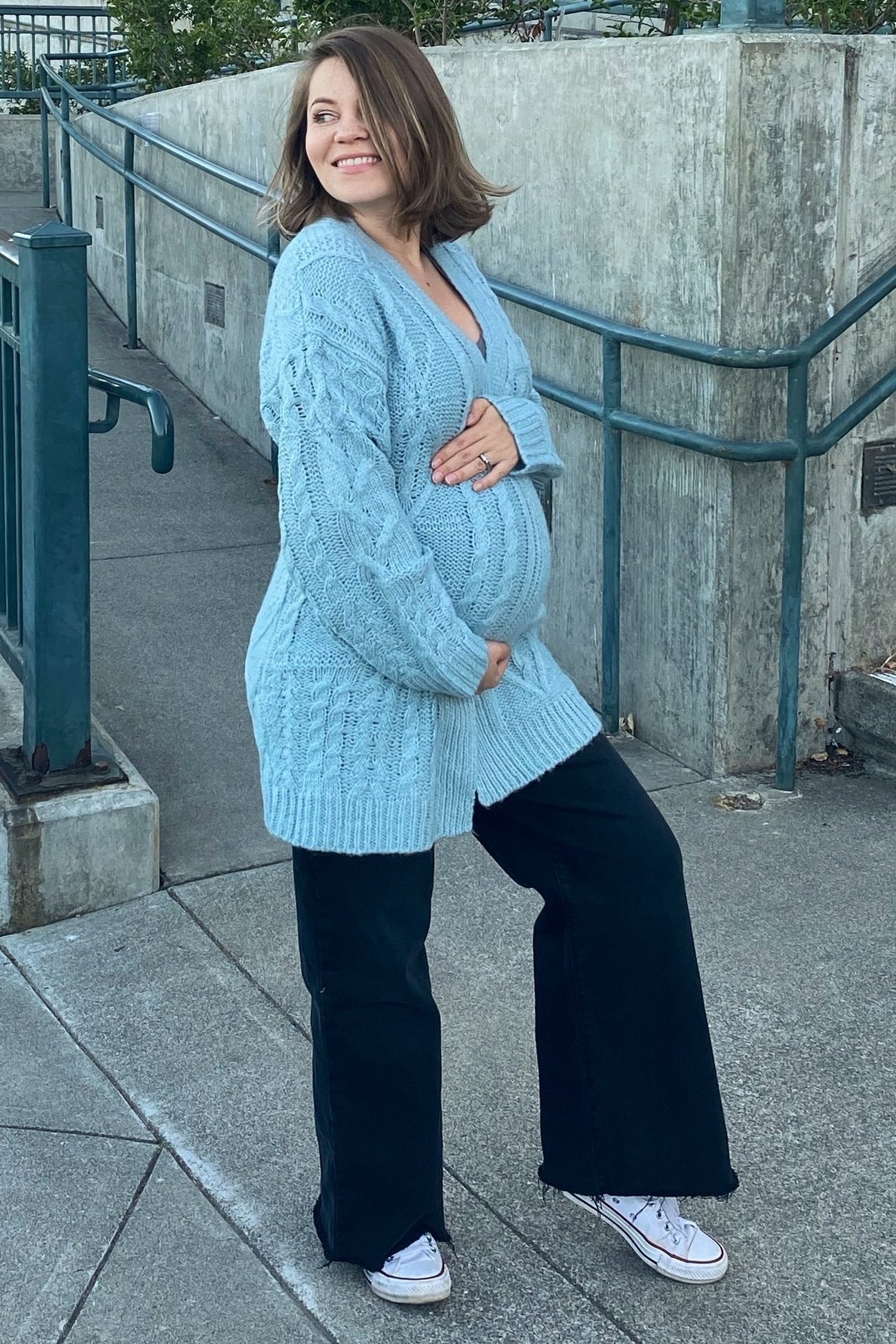 woman wearing a light blue knitted maternity cardigan and black wide-leg pants, holding her baby bump