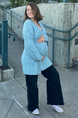 woman wearing a light blue knitted maternity cardigan and black wide-leg pants, holding her baby bump