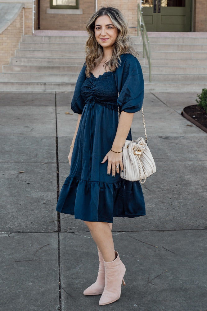 woman wearing a navy blue smocked dress with puff sleeves and a tiered skirt, holding a cream handbag and wearing pink ankle boots
