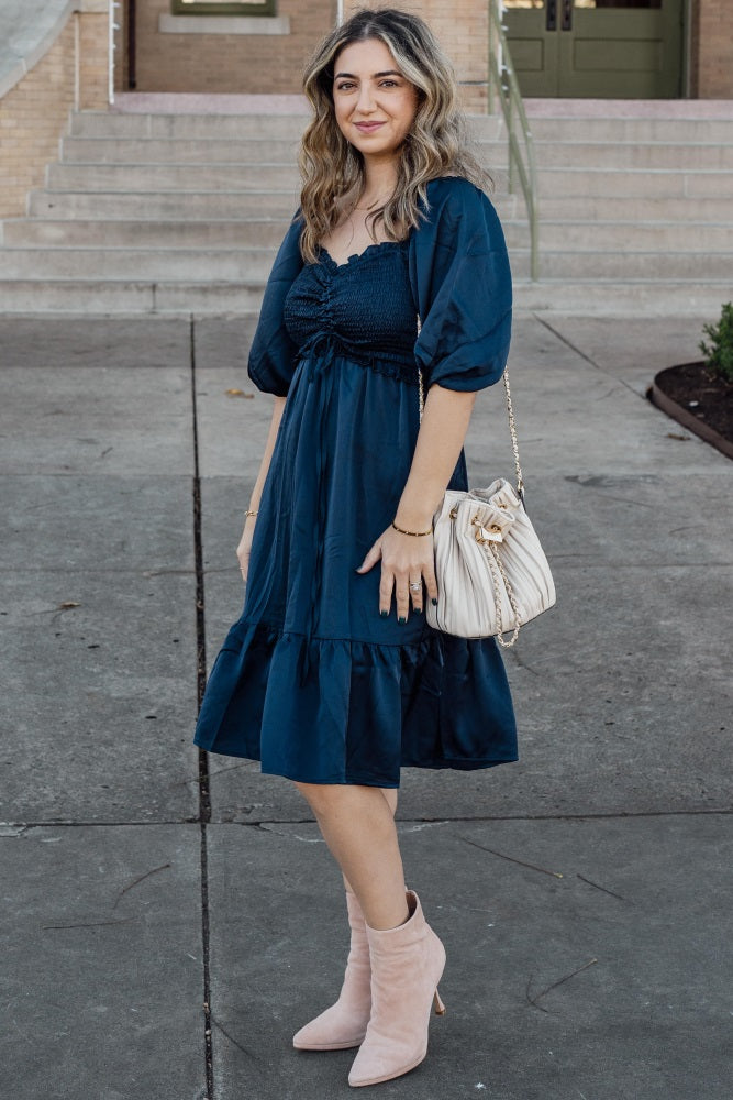 woman wearing a dark blue smocked bodice dress with puff sleeves and ruffled hem, holding a light bag, and wearing pink ankle boots