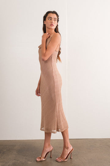 woman wearing a beige sleeveless midi dress and clear high-heeled sandals, posed sideways against a plain background