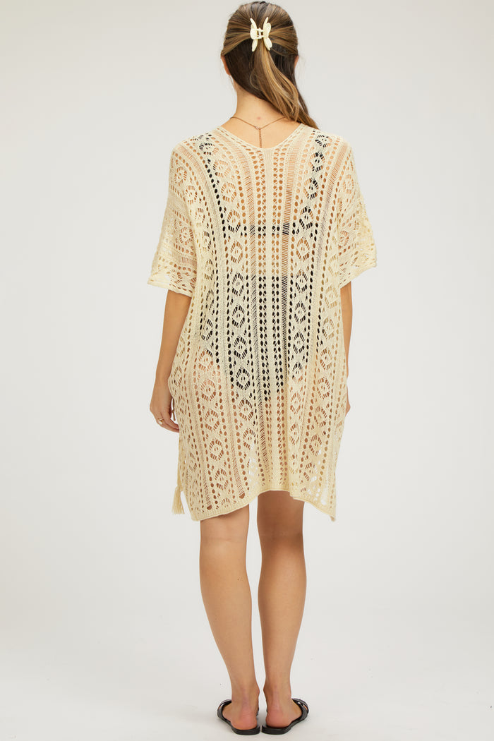 woman wearing a cream crochet maternity cover-up dress with a loose fit and short sleeves, shown from the back