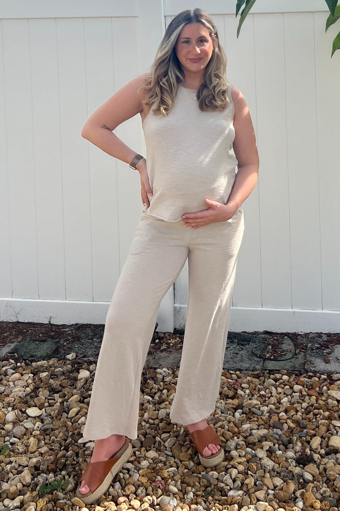 woman wearing a beige sleeveless maternity top and matching wide-leg maternity pants, gently cradling her baby bump