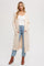 woman wearing a long beige cardigan over a white v-neck top and blue high-waisted jeans, standing with hands in pockets