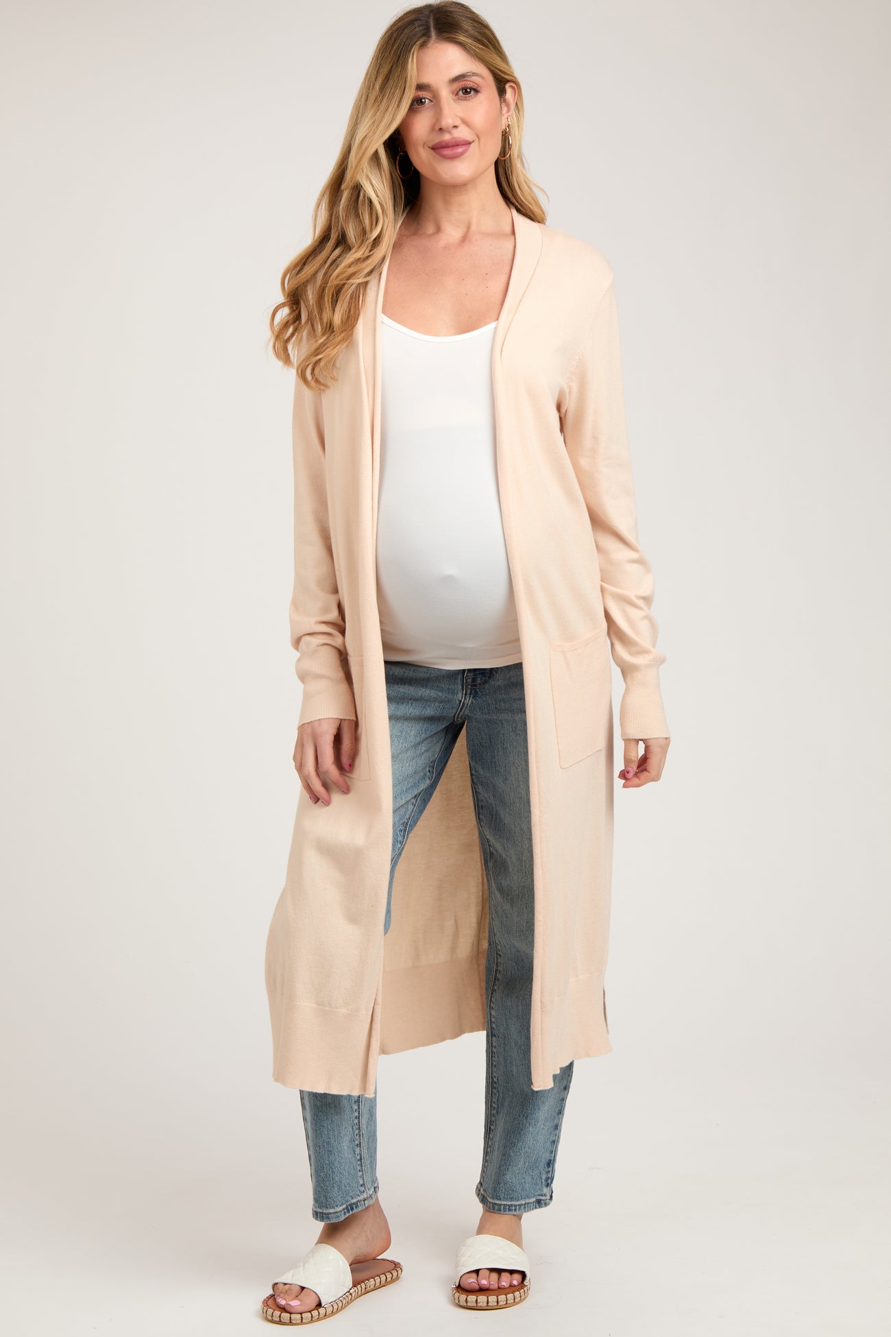 woman wearing a light pink maternity cardigan over a white top and blue jeans, showing her baby bump