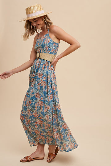woman wearing a blue floral maxi dress with a belted waist and a straw hat, standing and smiling