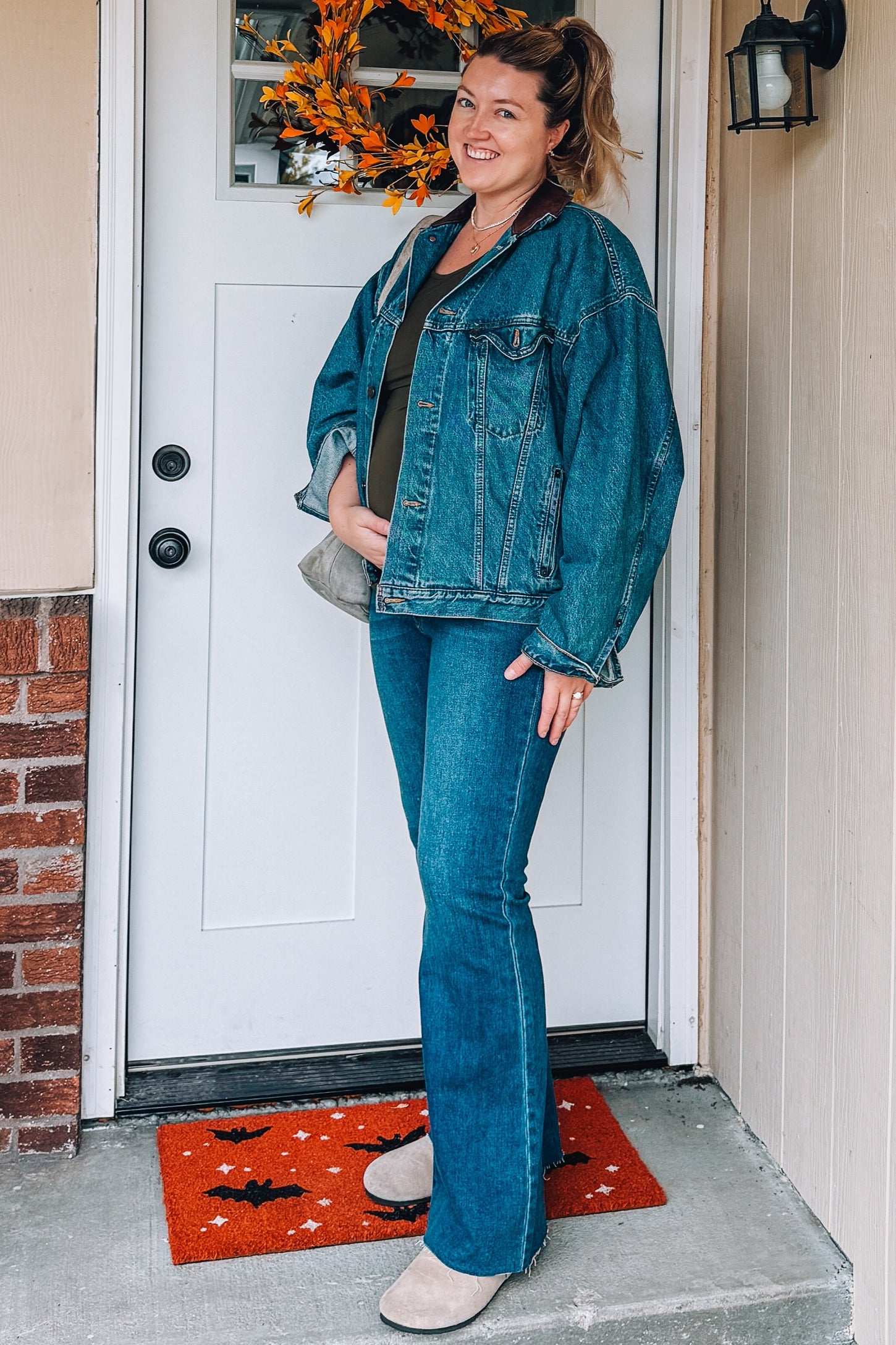 woman wearing a green maternity top, oversized denim jacket, and flared jeans, showing baby bump at the front door
