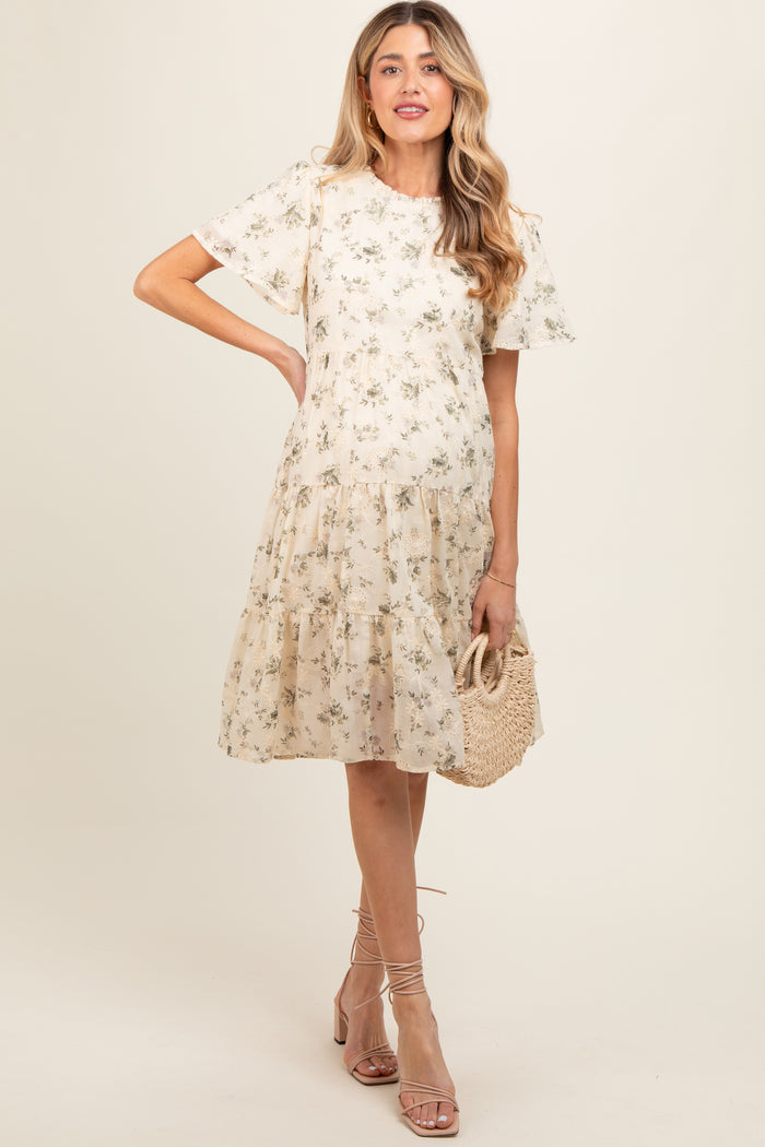 woman wearing a floral print beige maternity dress with short sleeves and a tiered skirt, holding a woven bag