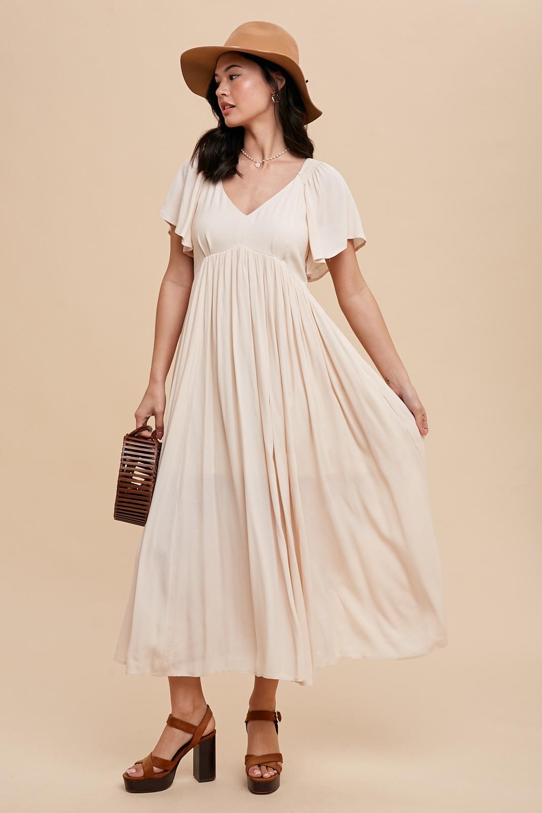 woman wearing a beige maxi dress with flutter sleeves and a v-neck, holding a brown handbag and wearing a wide-brimmed hat