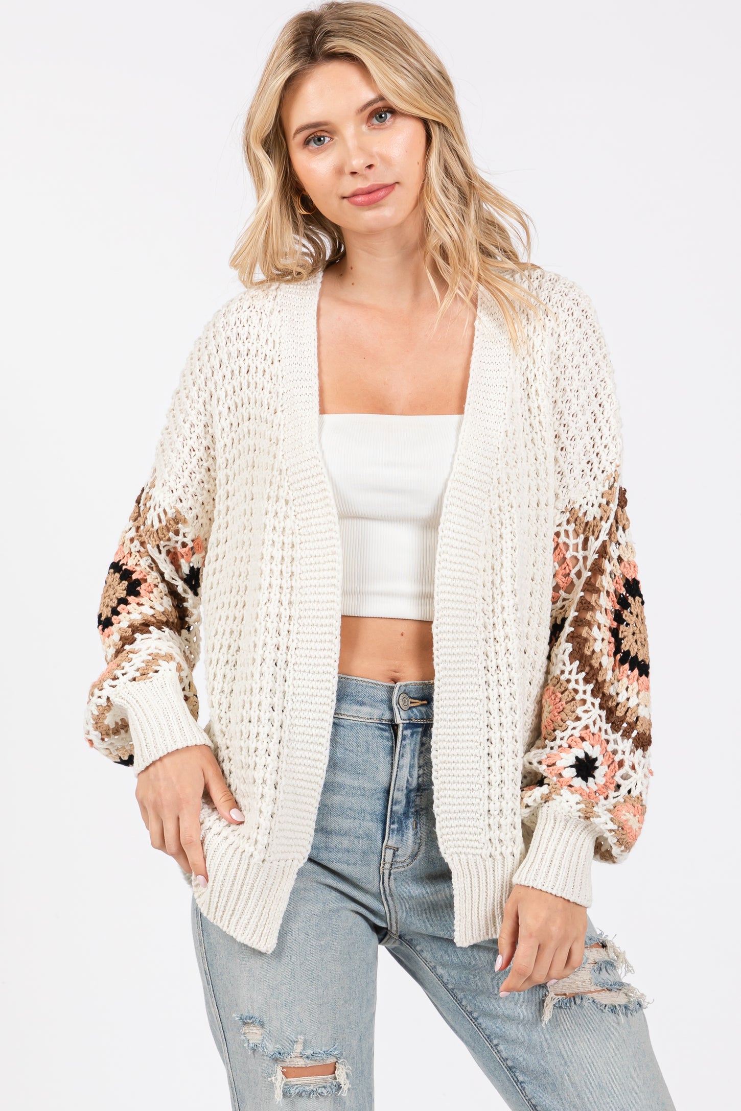 woman wearing a cream oversized crochet cardigan with colorful patterned sleeves and distressed light blue jeans