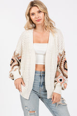 woman wearing a cream oversized crochet cardigan with colorful patterned sleeves and distressed light blue jeans