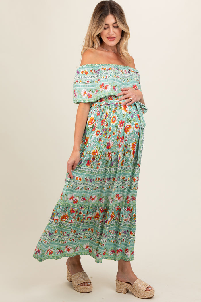 woman wearing a green floral off-the-shoulder maternity maxi dress and holding her baby bump